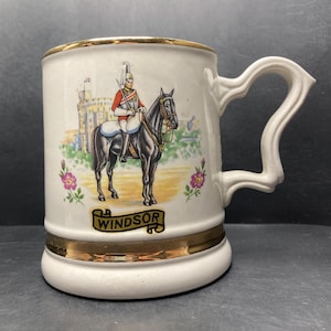 May include: White ceramic mug with a gold rim and a gold band around the bottom. The mug features a colorful illustration of a soldier on horseback in front of Windsor Castle. The text "Windsor" is written in gold on a banner.