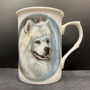 May include: White ceramic mug featuring a portrait of a fluffy white dog within a blue oval. The dog has a black nose and is smiling. The mug has a unique handle design and the artist's signature, "Sally Nash", is visible.