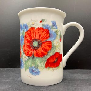 Op de afbeelding: Witte keramische mok met een bloemenontwerp. Het heeft grote rode klaprozen, blauwe korenbloemen en kleine witte madeliefjes. Het woord "Grace" staat op de zijkant gedrukt. De mok heeft een gebogen handvat.