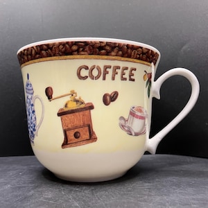 Pode incluir: Caneca de café de cerâmica creme com ilustrações de grãos de café, um moedor e a palavra "COFFEE". A borda é decorada com grãos de café. A caneca tem uma alça grande.