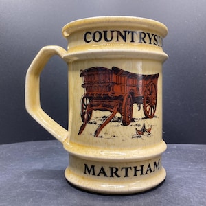 Może przedstawiać: Beżowy ceramiczny kubek z uchwytem, z brązową ilustracją wozu konnego. Na kubku widnieją napisy "COUNTRYSIDE" i "MARTHAM". Kubek jest w stylu vintage.