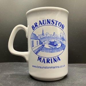 May include: White ceramic mug with a blue illustration of Braunston Marina, including a bridge and buildings. The mug has a curved handle and the words "Braunston Marina" are printed on the front, along with the website address.