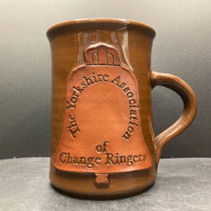 Op de afbeelding: Bruine keramische mok met een handvat. De mok heeft een verhoogd klokontwerp met de tekst "The Yorkshire Association of Change Ringers" in een cirkelvormig patroon rond de klok. De mok heeft een glanzende afwerking.