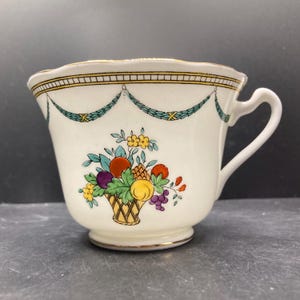 May include: Cream-colored teacup with a decorative fruit basket design. The cup features a gold rim and handle, with a scalloped edge. The fruit basket includes oranges, grapes, and other colorful fruits. The cup has a classic, vintage aesthetic.