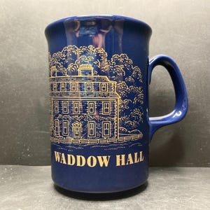 May include: A navy blue ceramic mug featuring a gold illustration of a building and the text "WADDOW HALL". The mug has a curved handle and a cylindrical shape. The building illustration includes windows and trees.