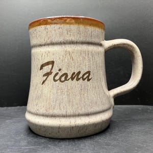 May include: A beige ceramic mug with a brown rim and handle. The name "Fiona" is etched in a cursive font on the front of the mug. The mug has a textured surface and a classic, sturdy design.