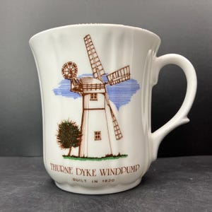 Op de afbeelding: Witte keramische mok met een bruine en witte illustratie van een windmolen en blauwe hemeldetails. De mok heeft de tekst "THURNE DYKE WINDPUMP BUILT IN 1820".