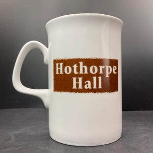 May include: White ceramic mug with a curved handle. The mug features the text "Hothorpe Hall" in white letters on a brown rectangular background. The mug is designed for hot beverages.