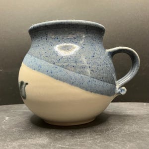 Könnte beinhalten: Eine handgefertigte Keramiktasse mit rundem Boden und blau gesprenkelter Oberseite. Die Tasse hat einen weißen unteren Teil und einen gebogenen Henkel. Auf der Seite ist ein kleines, dunkelblaues Design zu sehen.