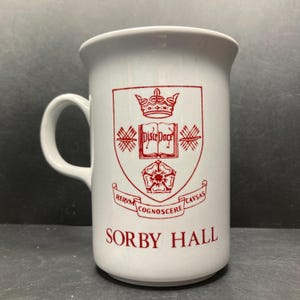 May include: White ceramic mug with a red crest design. The crest includes a crown, open book, and floral elements, with the words "Sorby Hall" at the bottom. The mug has a curved handle and a slightly flared rim.