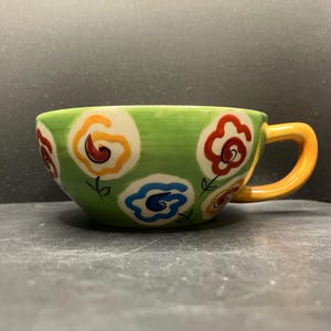 May include: A ceramic teacup with a green exterior, adorned with colorful floral designs in red, yellow, and blue. The cup has a yellow handle and sits on a dark surface. The floral pattern is repeated around the cup.