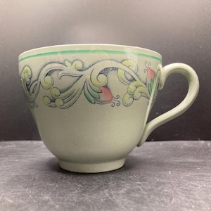 May include: A light green teacup with a curved handle and a decorative floral pattern in green, pink, and blue. The cup has a green band around the rim.