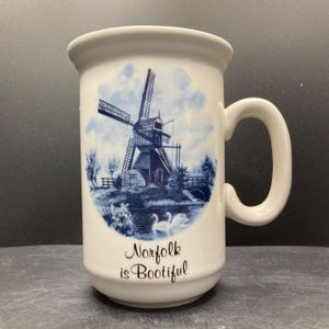 May include: White ceramic mug with a blue and white windmill scene, a bridge, and swans. The mug has a curved handle and the text "Norfolk is Bootiful" in a script font.
