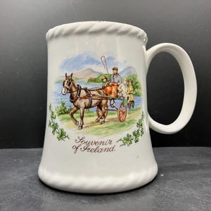 Op de afbeelding: Witte keramische mok met een handvat, met een kleurrijke illustratie van een paardenkar met mensen, tegen een schilderachtige achtergrond. De mok is versierd met de tekst "Souvenir of Ireland" en klaveraccenten.