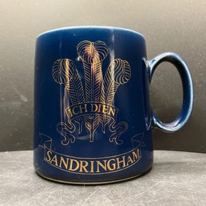 May include: A navy blue ceramic mug with a gold emblem featuring three feathers and the words "ICH DIEN" and "SANDRINGHAM". The mug has a curved handle and a glossy finish, suitable for hot beverages.
