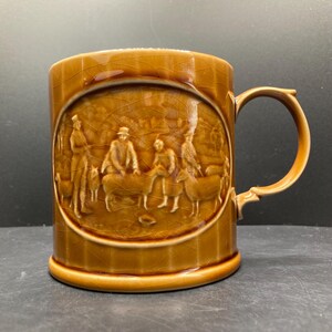 May include: A brown ceramic mug with a detailed relief scene of people and animals. The mug has a curved handle and a glossy finish. The relief depicts figures in a pastoral setting, adding a vintage aesthetic. The mug is a classic piece of drinkware.