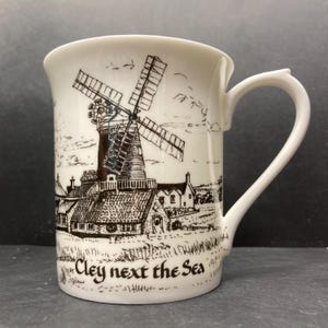 May include: White ceramic mug featuring a black and white illustration of a windmill and buildings. The mug has a curved handle and the text "Cley next the Sea" is printed on the front.