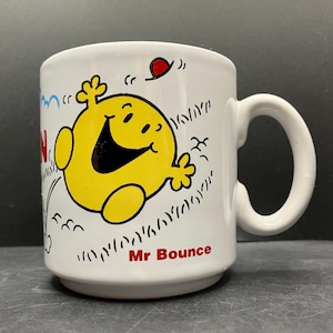 May include: White ceramic mug with a cartoon illustration of Mr. Bounce, a yellow character with a large smile. The mug has a curved handle and the words "Mr Bounce" in red.