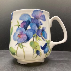 May include: White ceramic mug decorated with a watercolour design of blue and purple pansies and green leaves. The mug has a curved handle and a flared base. The floral design wraps around the mug.