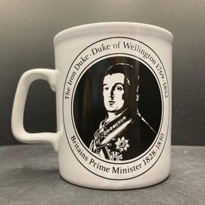 May include: White ceramic mug featuring a black and white portrait of the Duke of Wellington. The mug has a handle and text that reads "The Iron Duke - Duke of Wellington 1769-1852, Britains Prime Minister 1828-1830."