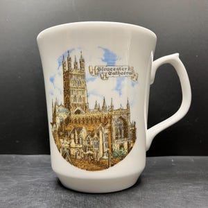 May include: White ceramic mug featuring a detailed illustration of Gloucester Cathedral. The design includes the cathedral's architecture, blue sky, and the text "Gloucester Cathedral". The mug has a curved handle.