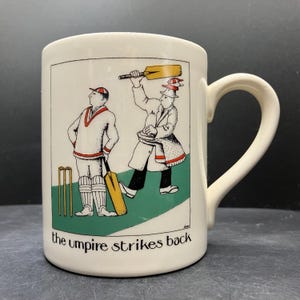 May include: Cream ceramic mug with a curved handle. The mug features a cartoon illustration of cricket players and the text "the umpire strikes back". The illustration includes a batsman, umpire, and cricket equipment.