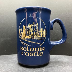 May include: A navy blue ceramic mug with a gold illustration of Belvoir Castle. The castle image is within a circle, and the words "Belvoir Castle" are printed below. The mug has a curved handle.