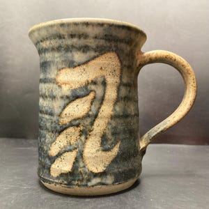 Op de afbeelding: Een handgemaakte keramische mok met een uniek ontwerp. De mok heeft een beige, gestileerd symbool op een achtergrond van blauwe en grijze horizontale strepen. De handgreep en de rand zijn beige. Perfect voor koffie of thee.