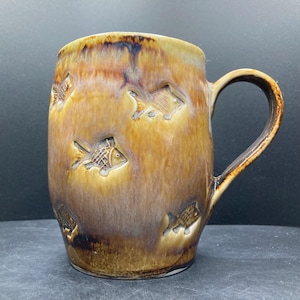 Könnte beinhalten: Eine handgefertigte Keramik-Tasse mit brauner und goldener Glasur. Die Tasse hat geprägte Fischmotive am Körper. Der Griff hat eine passende braune Farbe. Die Tasse ist für heiße oder kalte Getränke geeignet.