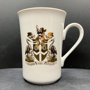 May include: White ceramic mug with a gold crest design. The crest includes two herons, a shield, and the words "FOR HOME AND COUNTRY." The mug has a curved handle and a flared rim. The design is centered on the mug.