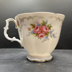 Peut inclure: Tasse à thé crème avec un motif floral de roses roses et d'accents bleus. La tasse a une anse décorative et une bordure dorée. La tasse à thé est probablement en porcelaine.