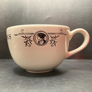 May include: Cream ceramic teacup with a large handle. The cup has a black circular design with a woman's profile and the words "Charlotte Watson". Decorative black flourishes and text are also present.