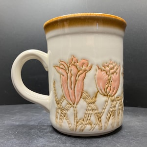 May include: Cream-colored ceramic mug with a brown rim and handle. The mug features embossed pink tulip designs with brown stems and leaves. A vintage-style coffee or tea cup.