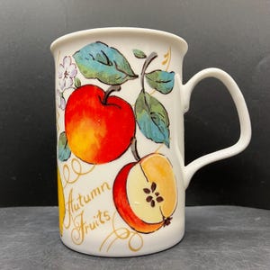 以下が含まれることがあります： 白いセラミックマグカップ、ハンドル付き、リンゴ、葉、そして「Autumn Fruits」のカラフルなイラストで装飾されています。リンゴは赤と黄色で、緑と青の葉があります。