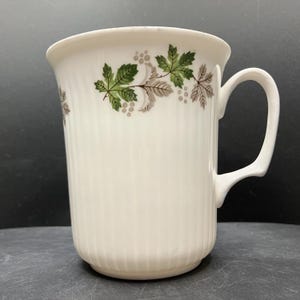 May include: White ceramic mug with a ribbed texture and a curved handle. The mug is decorated with a pattern of green and gray leaves and berries around the rim. The mug is designed for hot beverages.