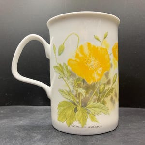 May include: White ceramic mug with a yellow poppy floral design and green leaves and buds. The mug has a curved handle and a flared rim. The artist's signature, "Rosine Girard," is visible near the base. A decorative piece for the kitchen, perfect for tea or coffee.