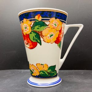 May include: A white ceramic mug with a floral design. The mug features a blue rim and base with a white handle. The floral pattern includes orange, yellow, and red flowers with green leaves. The mug is approximately 15 cm tall.