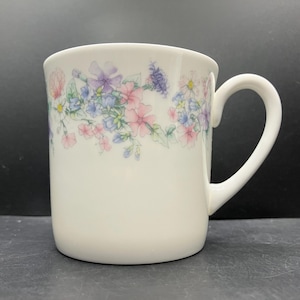 May include: White ceramic mug with a floral design featuring pink, purple, and blue flowers. The mug has a curved handle and a smooth, glossy finish. The floral pattern is arranged in a band around the top of the mug.