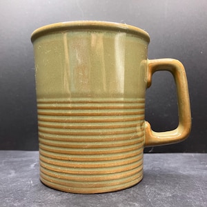 May include: A ceramic mug with a light green glaze and horizontal, textured bands. The handle and rim are a darker shade of green. The mug is cylindrical and appears to be sturdy.