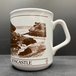 May include: White ceramic mug with a brown illustration of a coastal landscape. The mug features the word "BOSCASTLE" in brown text at the bottom. The mug has a curved handle and a simple, clean design.