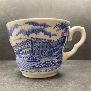 May include: A white ceramic teacup with a blue and white design of Hampton Court Palace in London. The cup has a handle and a flared rim.