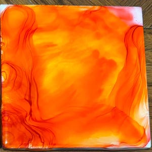 Op de afbeelding: Vierkant kunstwerk met een levendig abstract ontwerp. Het stuk heeft een mix van oranje en gele tinten, met donkerdere rood-oranje accenten die vloeiende, organische vormen creëren. Het kunstwerk heeft een glanzende afwerking.