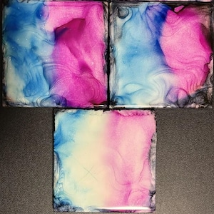 May include: Four square coasters with abstract designs. Each coaster features a blend of blue, pink, and white hues, creating a marbled effect. The edges are dark, framing the colorful patterns. These coasters are ideal for home decor.