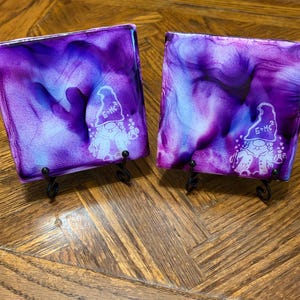 May include: Two square coasters featuring a vibrant purple, blue, and white alcohol ink swirl pattern. Each coaster displays a white line art graphic of a gnome wearing a hat with the text E=MC2 printed on it, supported by black metal stands.