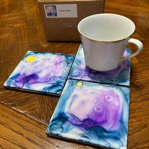 Pode incluir: Conjunto de quatro bases quadradas com um design celestial em tons de azul, roxo e branco. Uma chávena branca com uma borda dourada está em cima de uma base. A caixa no fundo tem o texto "Princess Asha".