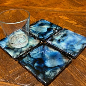 May include: Set of four blue and black abstract coasters with a swirling pattern. The coasters are made of resin and have a glossy finish.