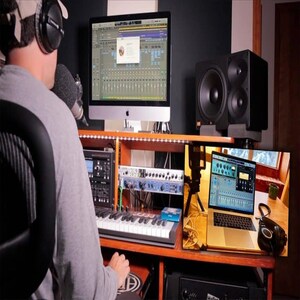 May include: A person sits in front of a computer with a microphone and headphones, working on a music production project. The computer screen shows a digital audio workstation (DAW) with multiple tracks and plugins. A keyboard, audio interface, and studio monitors are also visible.