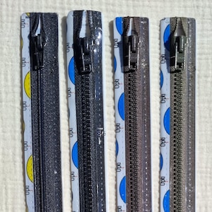 Op de afbeelding: Een verticale opname van vijf gesloten ritsen in verschillende tinten grijs en bruin. Elke rits heeft een metalen trekker. De ritstape heeft een herhalend patroon van gele en blauwe halve cirkels met het woord "ndo".