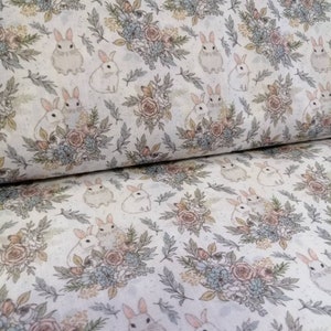 Op de afbeelding: Witte stof met een herhalend patroon van kleine witte konijntjes en bloemboeketten in tinten roze, blauw en groen.