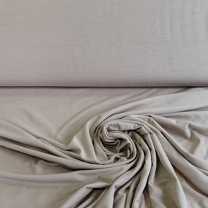 Może przedstawiać: Zbliżenie na miękką, jasnobrązową tkaninę o gładkiej fakturze. Tkanina jest udrapowana i złożona, prezentując swój drapowanie i przepływ.